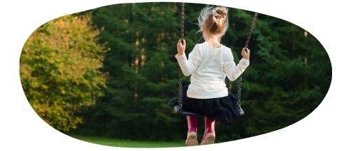 Girl on a swing with hair in a bun, white long sleeved top, navy skirt and pink wellies