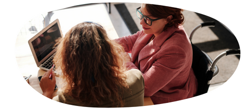 Girl with wavy brown hair talking to an adult in a pink jacket with glasses using a computer